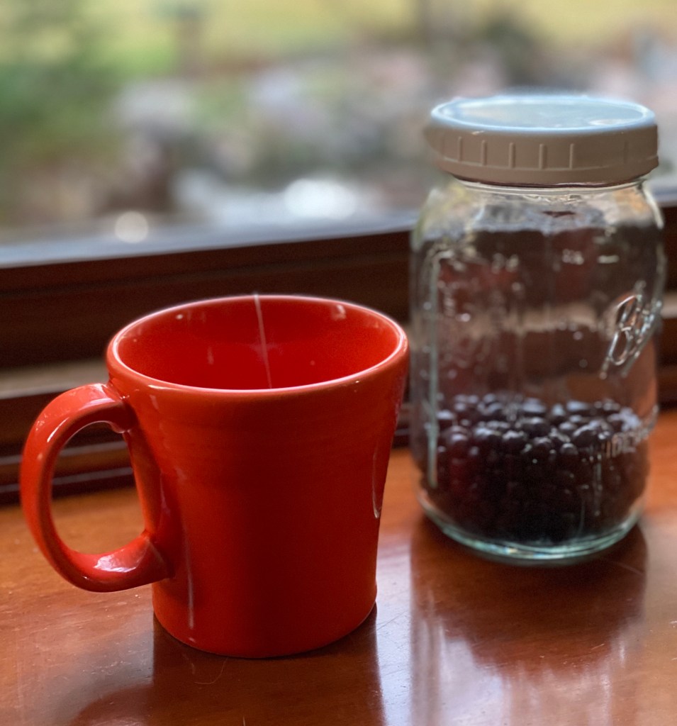 Brightly colored Orange stoneware tea mug with a glass "ball" canning jar of coffee beans. window behind both cup and jar of coffee beans. close up of mug and coffee beans