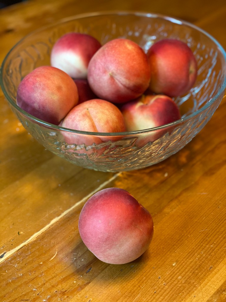 A pretty clear glass bowl with a decorative leaf pattern is filled with white peaches. Glass bowl is resting on a wooden table. A single peach is on the table closest to the  camera.